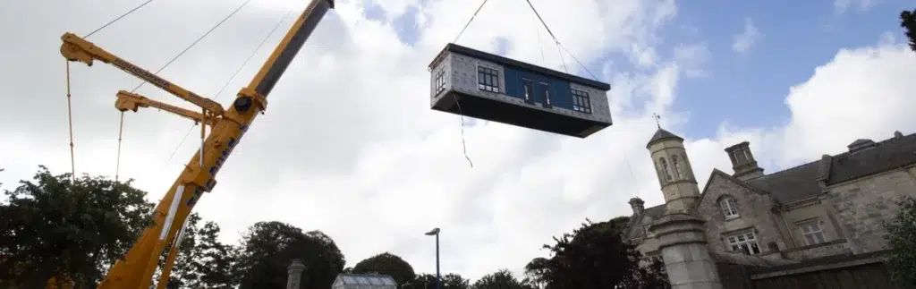 Crane lifting a grey modular building module into position at a historic site, demonstrating efficient value engineering construction methods