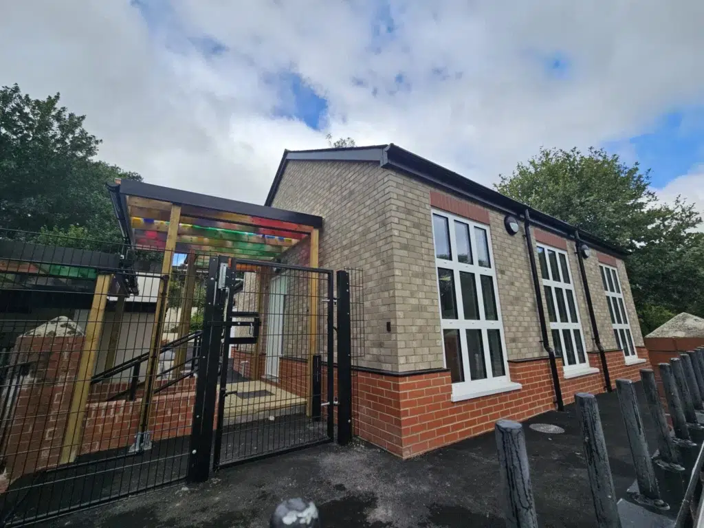 Side view of the new brick building at Cerne Abbas First School, featuring large windows and a colourful covered walkway leading to a gated entrance.