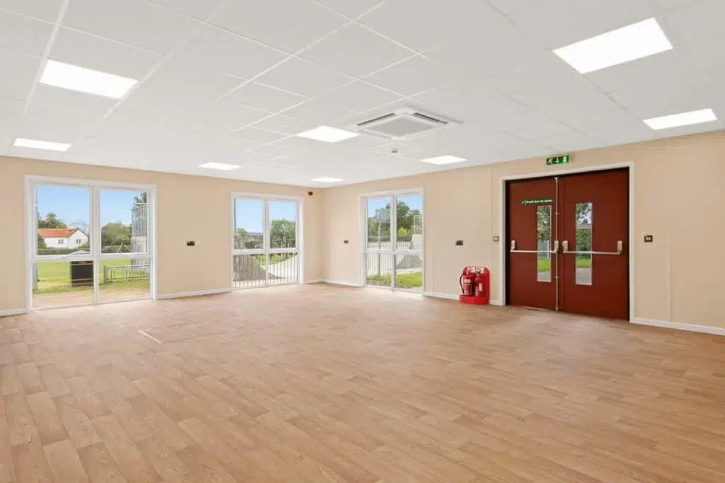 Modern modular community centre interior with flexible open-plan layout, wooden parquet flooring, large windows providing natural light and fire-rated doors