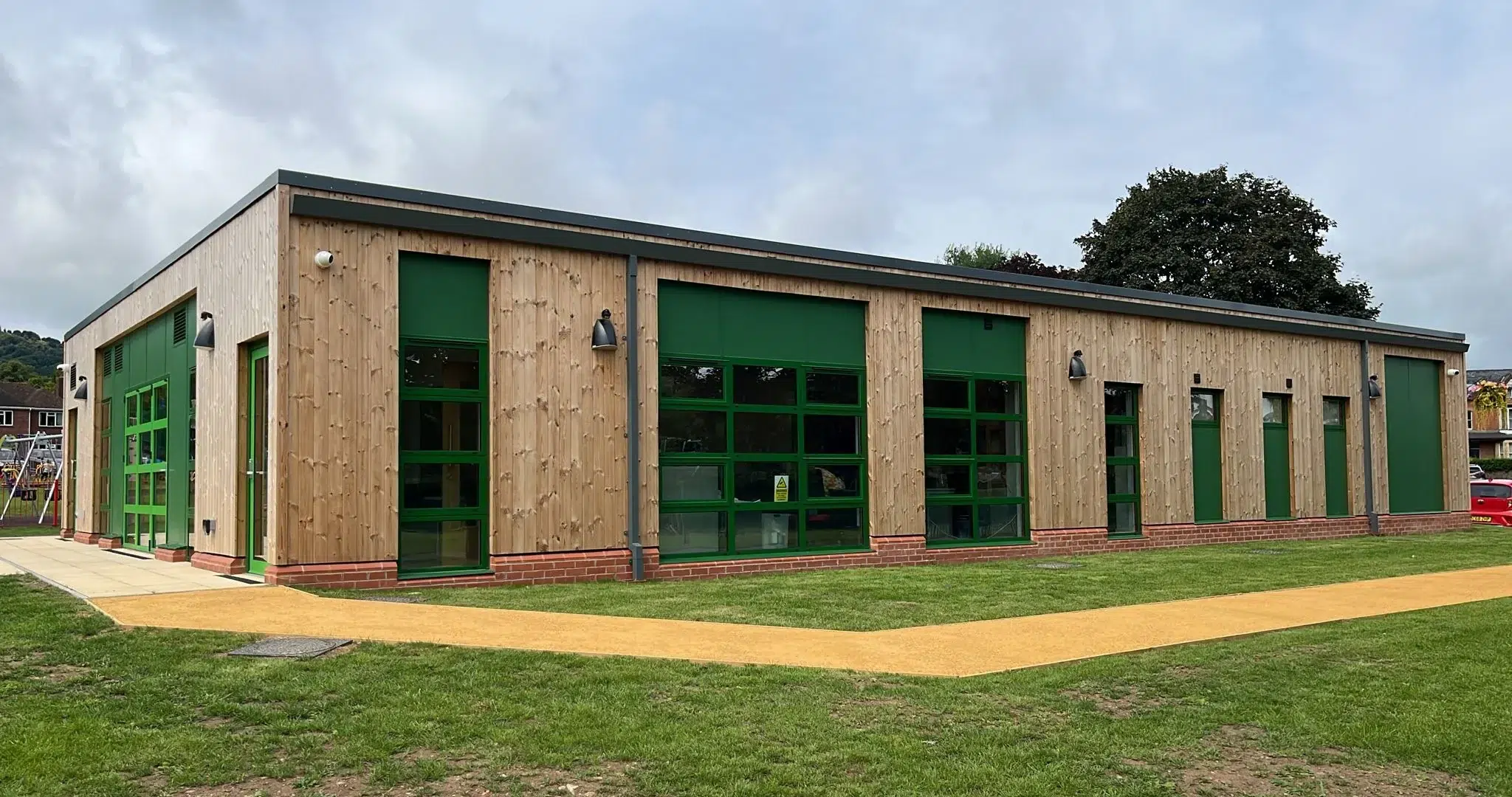 Modern timber-clad modular community centre featuring natural wood cladding, bright green window frames and doors, brick plinth base, and accessible yellow pathway for wheelchair users, set in playground area with trees